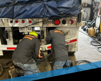 Two workers in safety vests crouch under a truck, repairing its underside in a garage