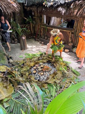 Person in floral dress leans over a firepit pointing to cooking food, with others nearby and greenery around.