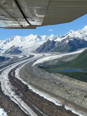 Glacier winding through snowy mountains beneath a blue sky, seen from under a plane wing