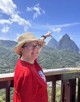 Person in red shirt and straw hat pointing at St. Lucia's Piton mountains under a bright blue sky.