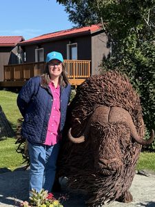 Woman standing beside a large shaggy brown Musk Ox sculpture outdoors near a house.