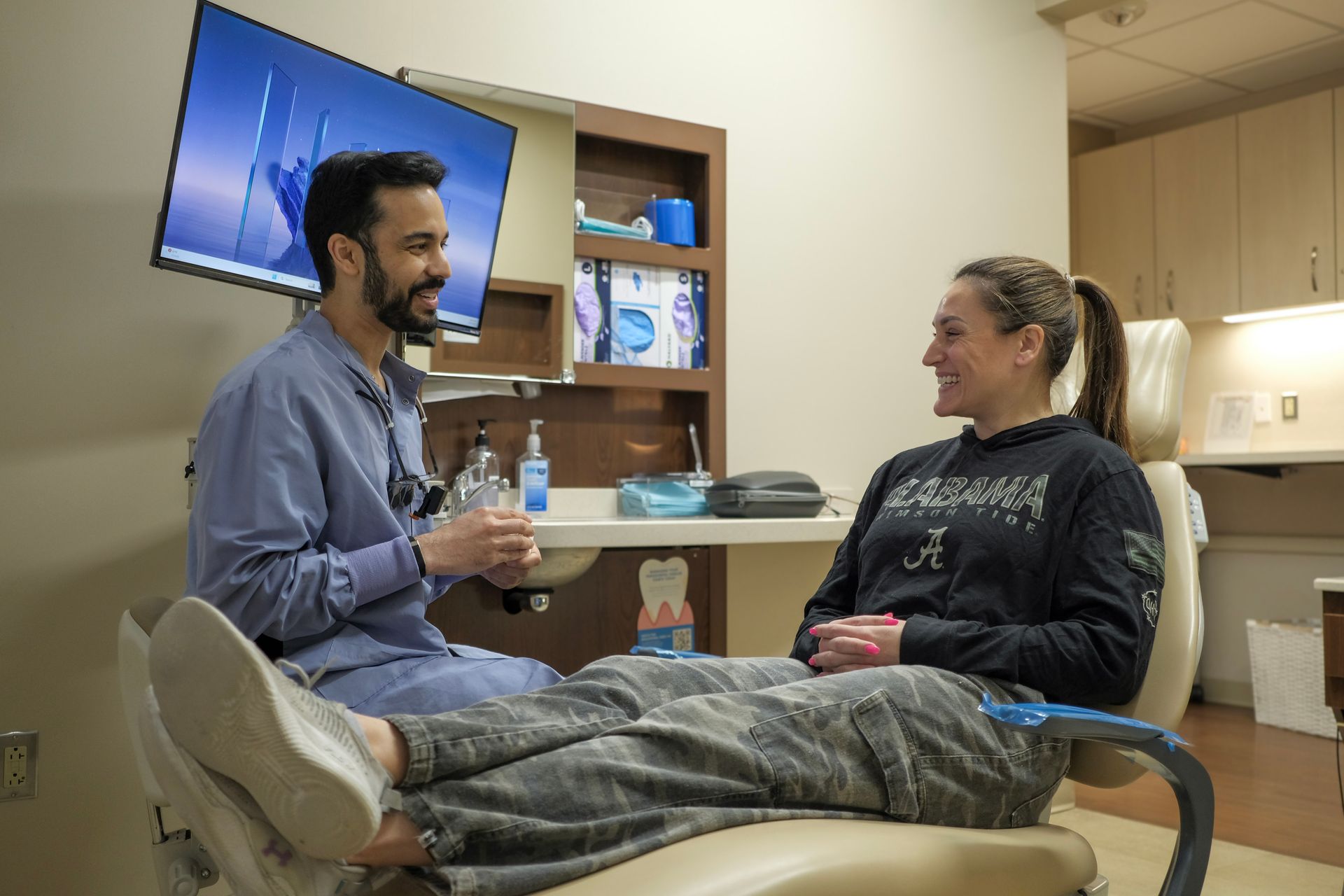 Clinician talks with seated patient in a dental exam room, with monitor and cabinets in the background