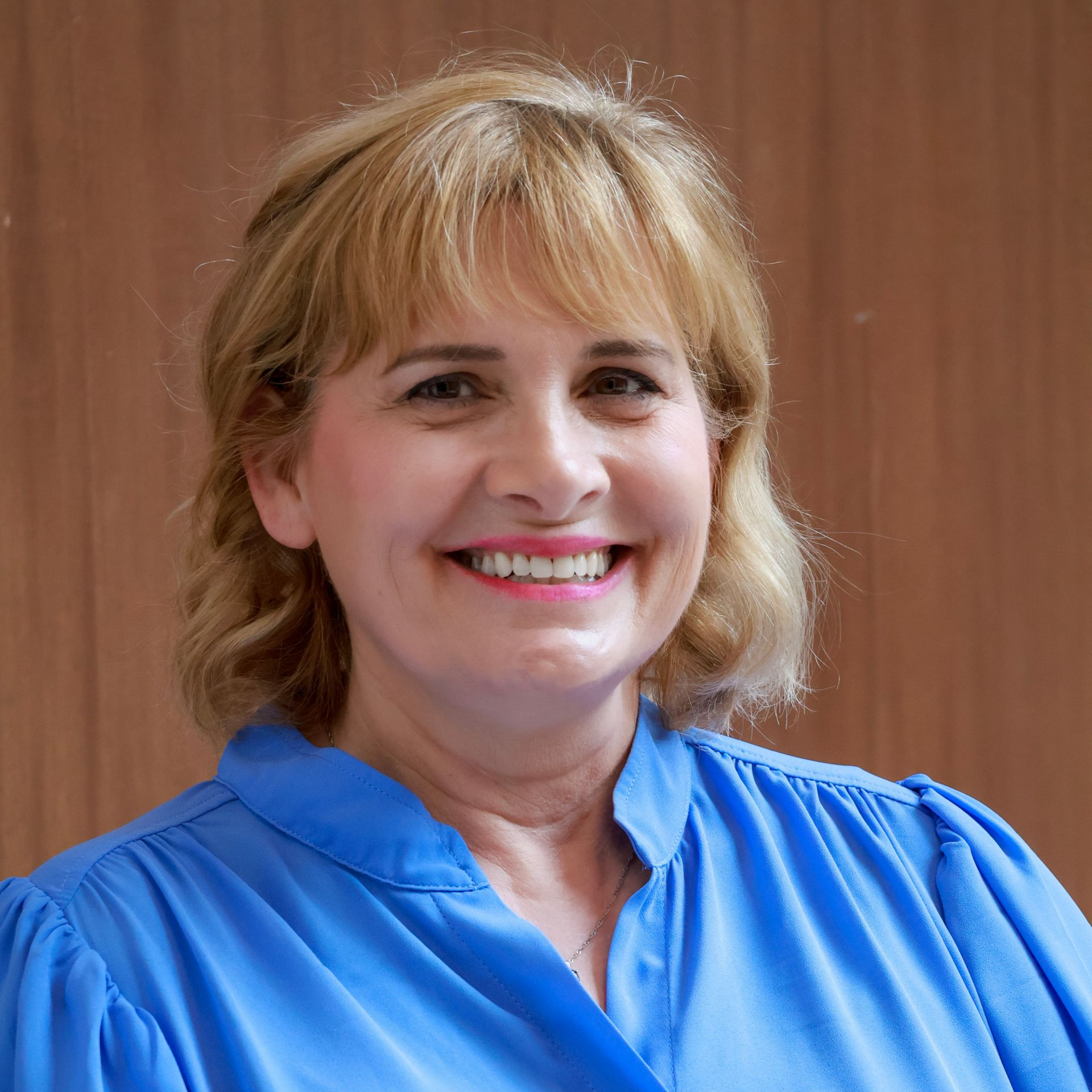 Woman in a white coat smiles, pink shirt visible, looking at the viewer.