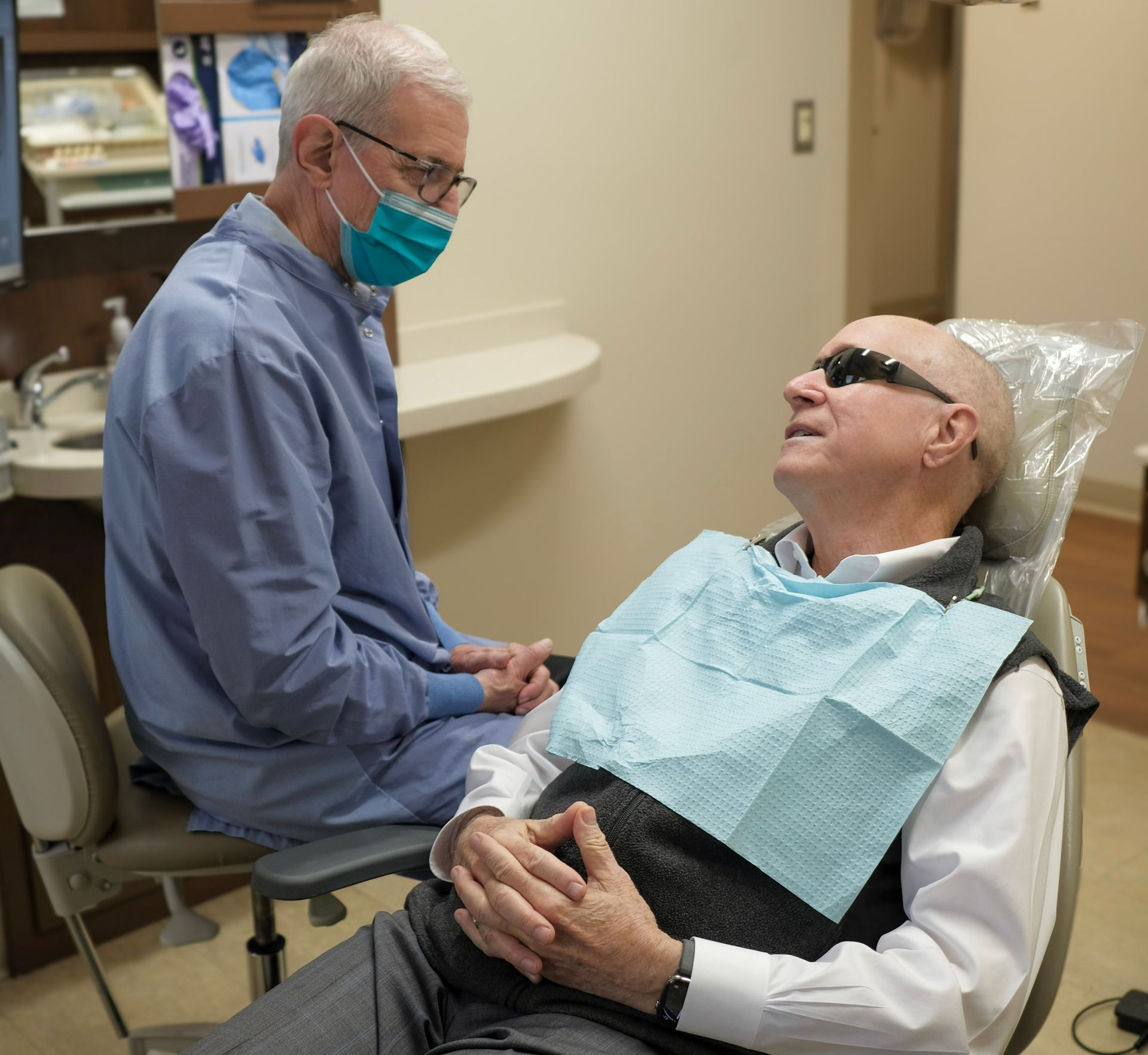 Woman in dental chair, grimacing and holding jaw, with a dentist and assistant nearby.