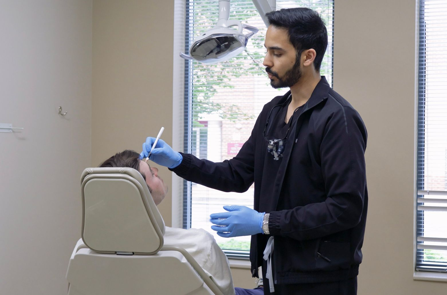 Dentist examining a patient's face in an office setting. Both are wearing masks and gloves.
