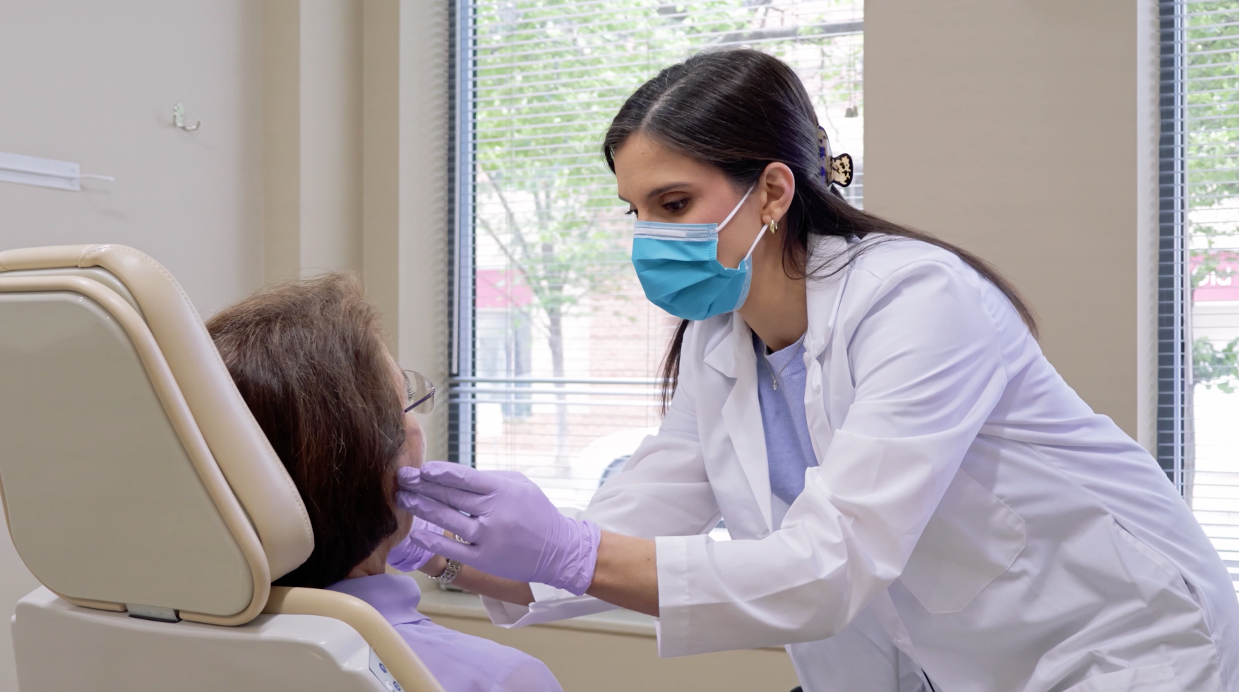 Woman in dentist chair smiles, touching her cheek. Dentist in mask is looking at her.