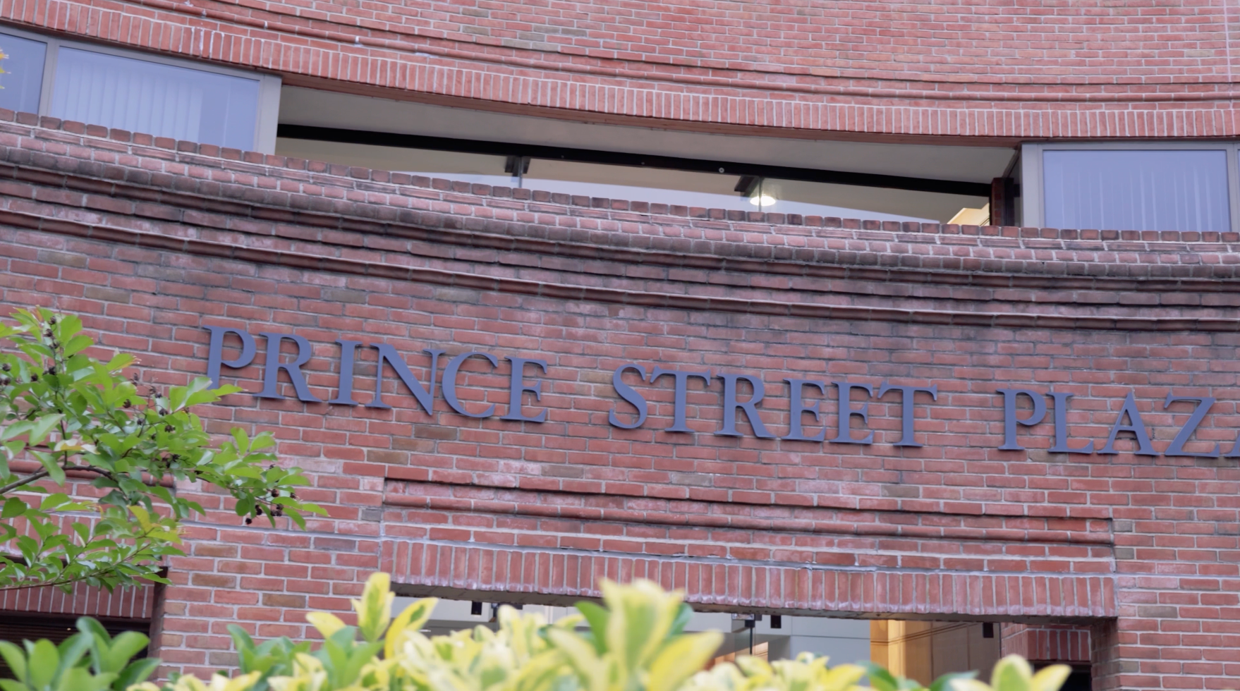Brick building sign reading “Prince Street Plaza” above greenery.