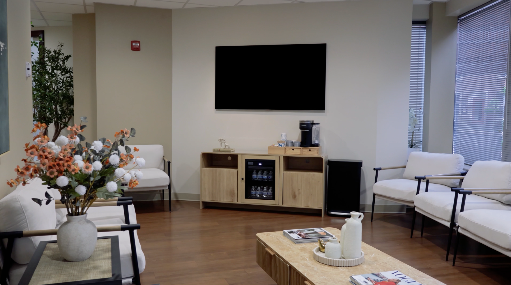 Modern waiting room with white sofas, coffee table, wall-mounted TV, and floral arrangement