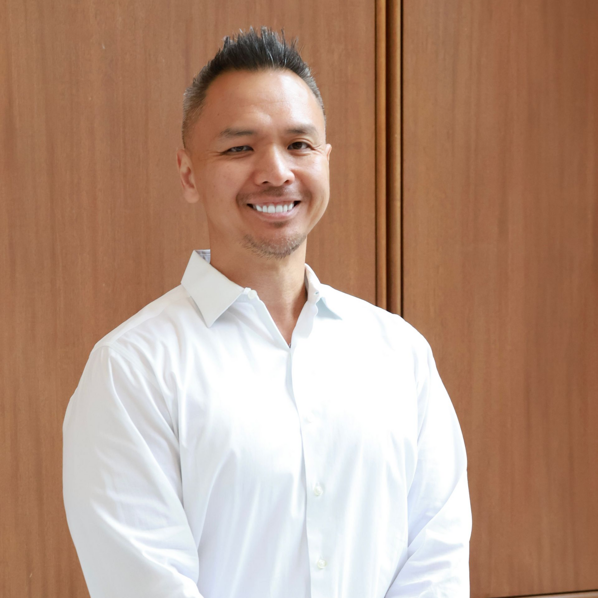 Man in suit smiles at the camera, light skin, short dark hair, blue shirt, dark tie, white background.