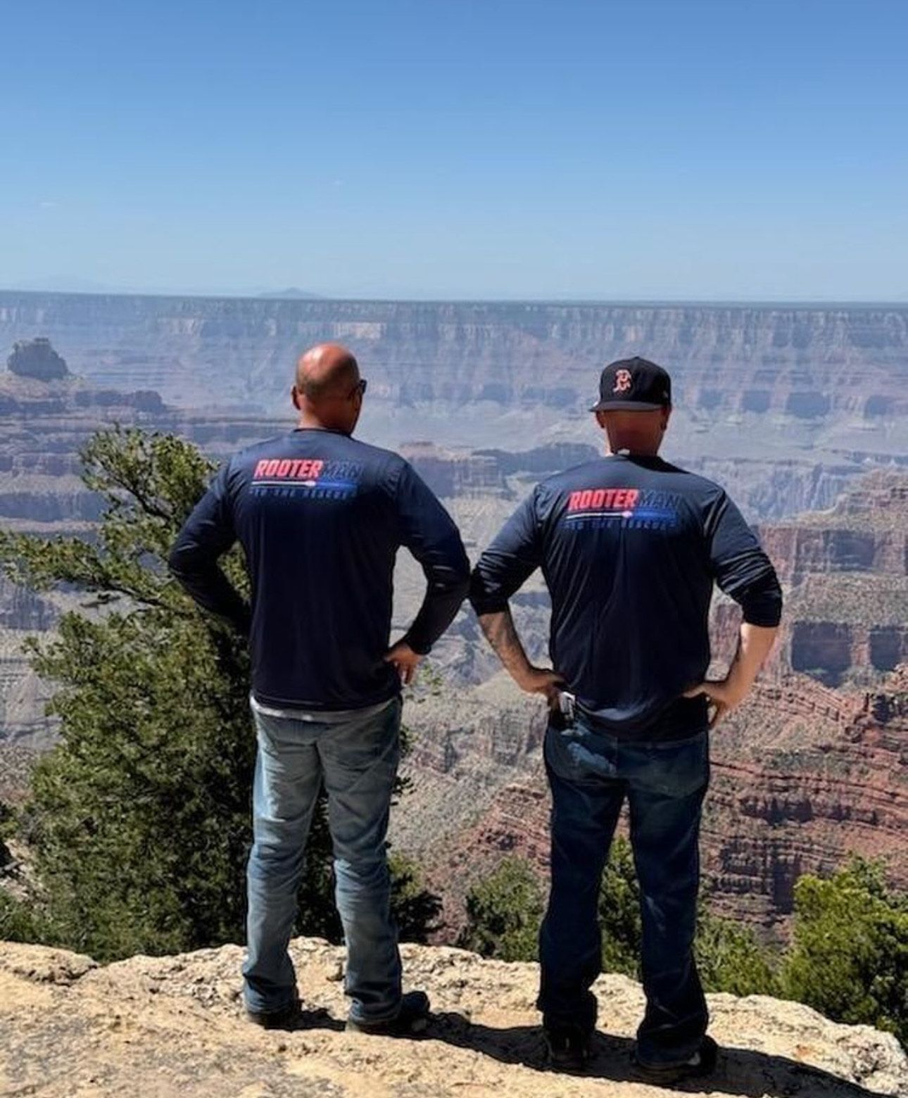 Two hikers in dark jackets overlook a vast canyon from a rocky cliff edge.