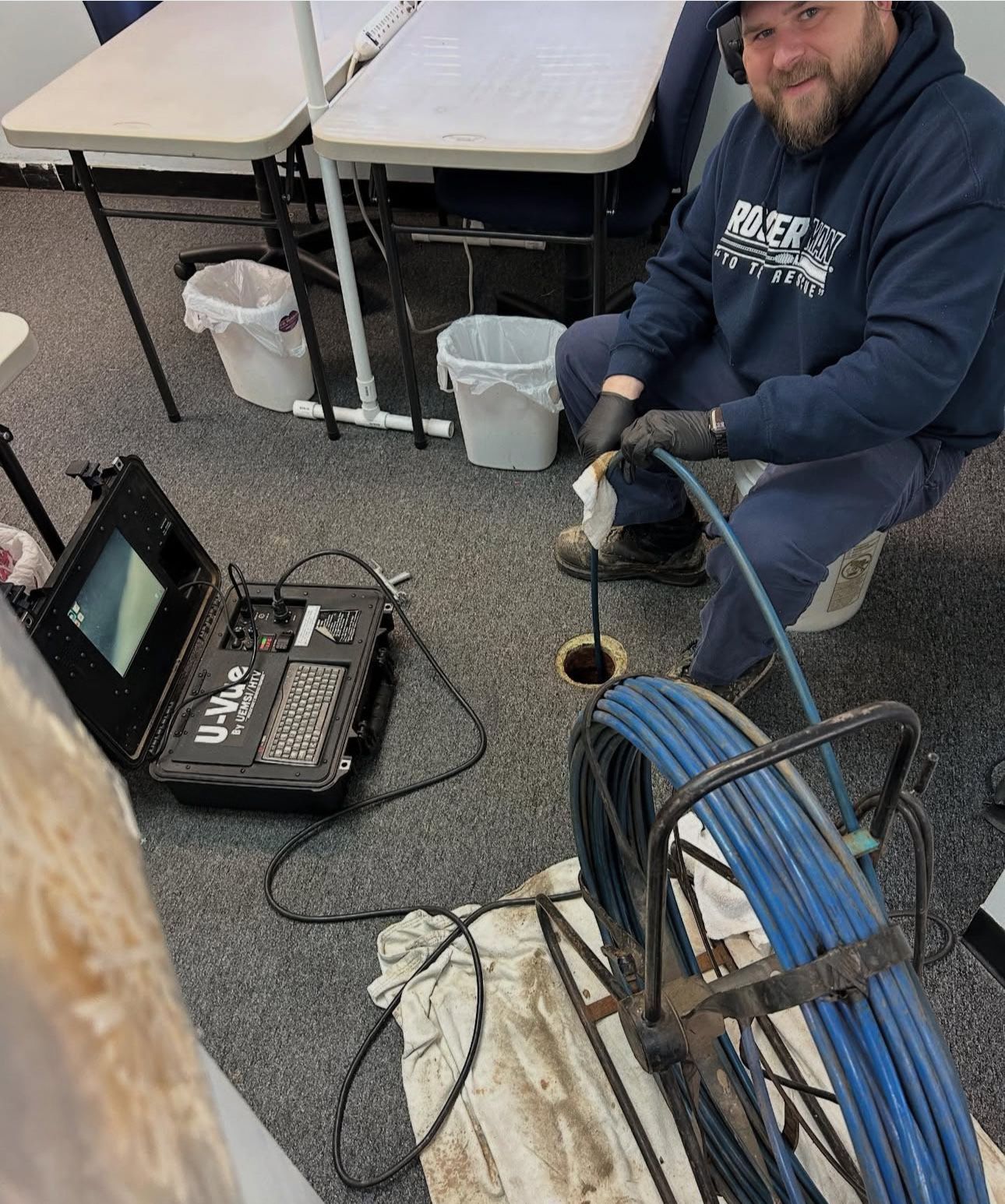 Man in a blue hoodie inspects a drain with a camera in an office setting. He's smiling.