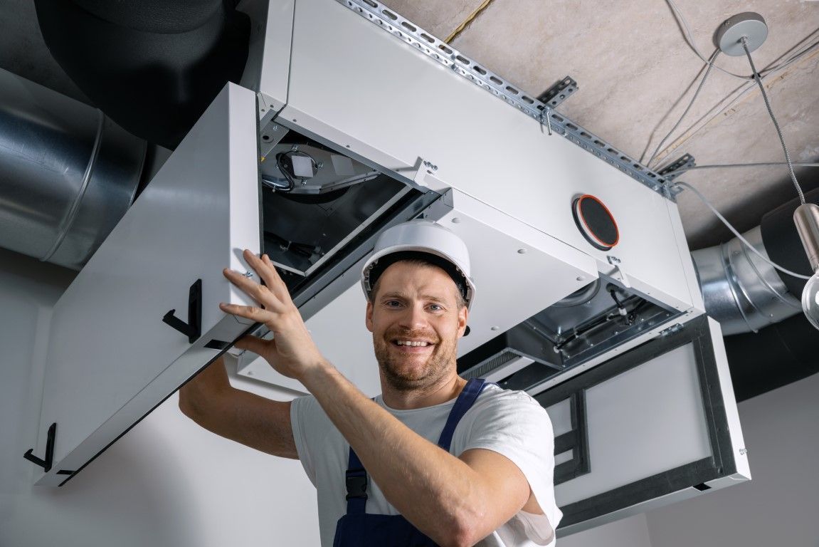 Technician opening an HVAC ceiling panel in a utility room, smiling and wearing a hard hat.