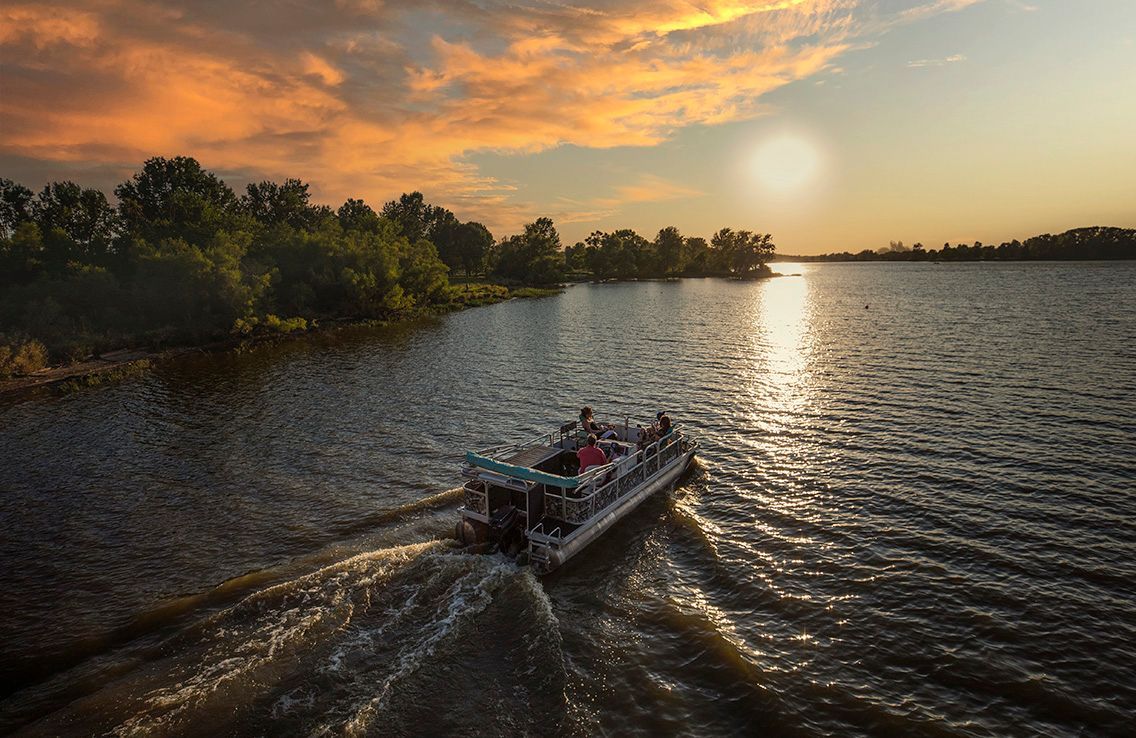 A pontoon boat cruises on a river at sunset, with a vibrant, warm sky and lush green trees along the shoreline.