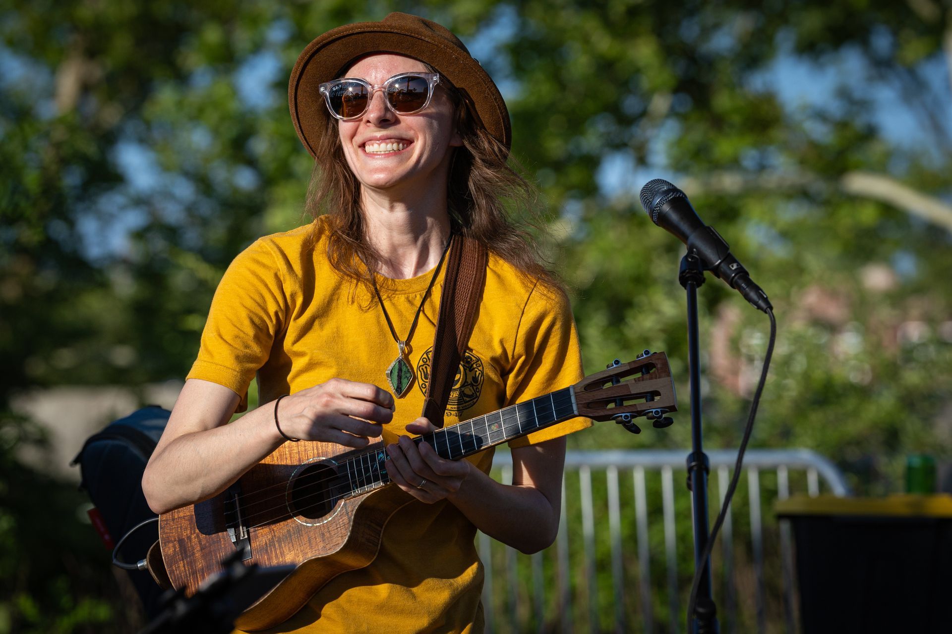 A smiling person wearing a hat and sunglasses plays a ukulele outdoors near a microphone stand.