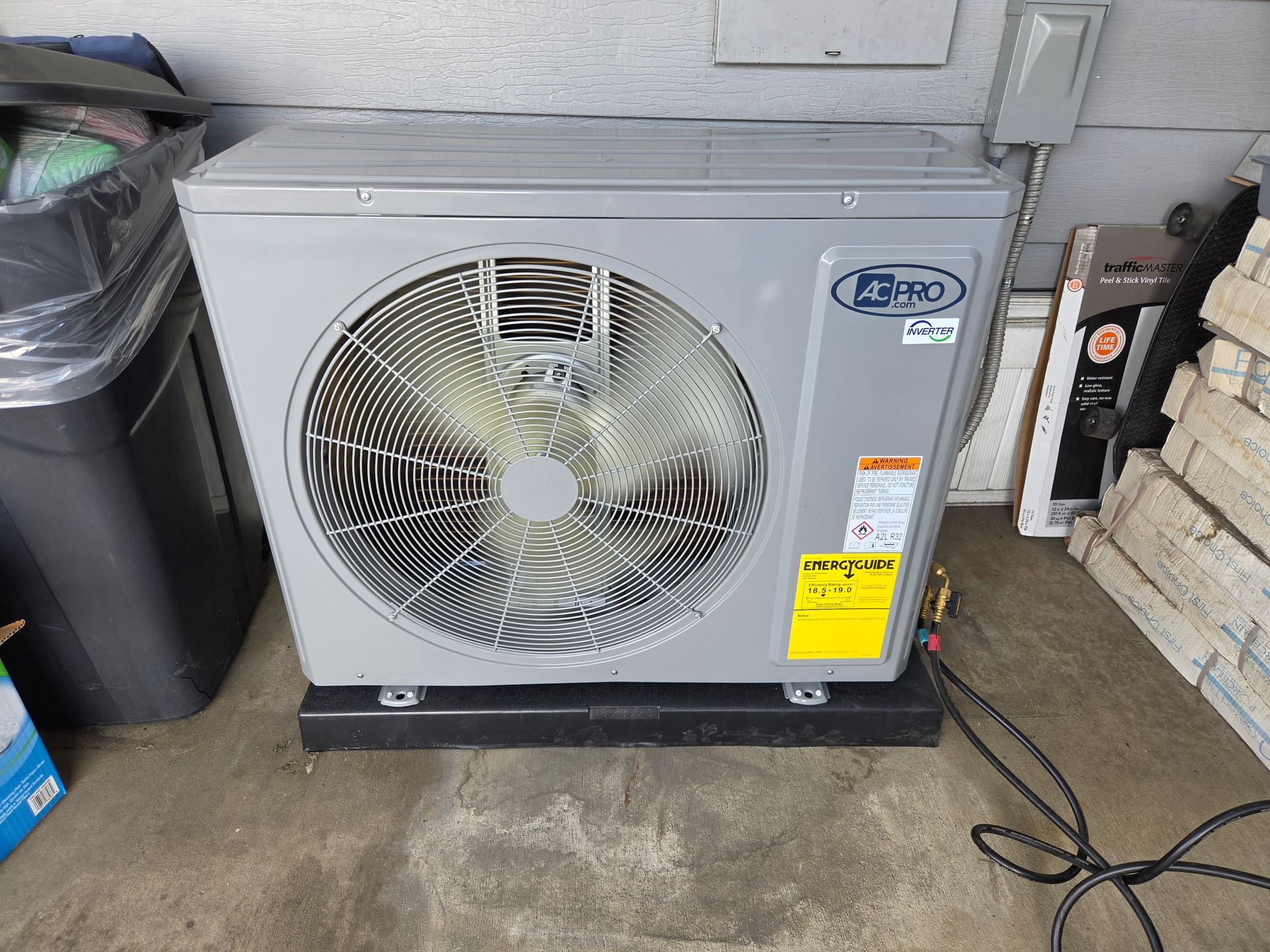 Outdoor air conditioner unit with large fan, sitting on a concrete floor beside a wall and trash bin.