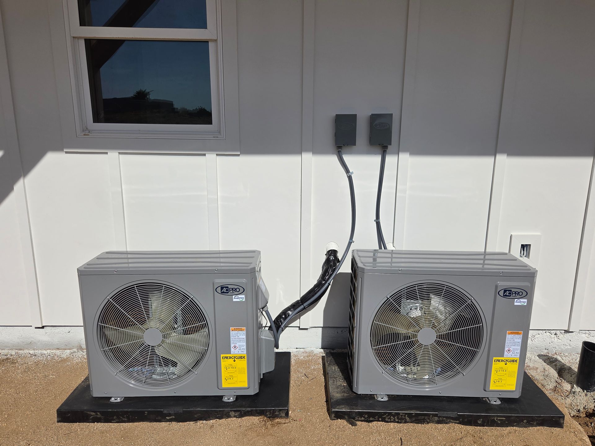 Two gray outdoor heat pump units on a concrete slab beside a white house wall and window.