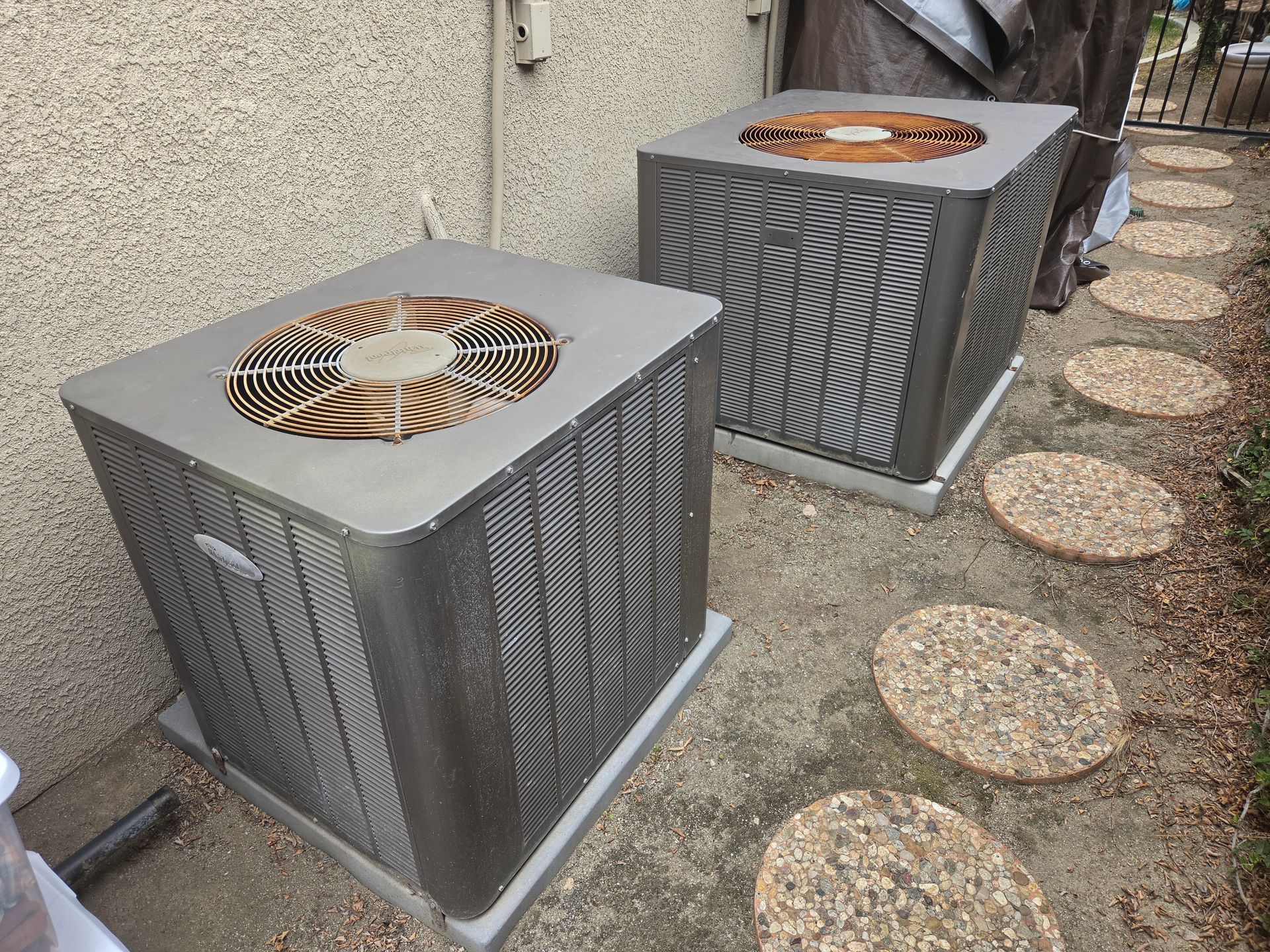 Two outdoor HVAC units beside a stucco wall and a gravel path with round stepping stones.