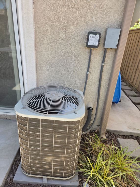 Outdoor air conditioner unit beside a stucco wall, with electrical boxes and a walkway nearby