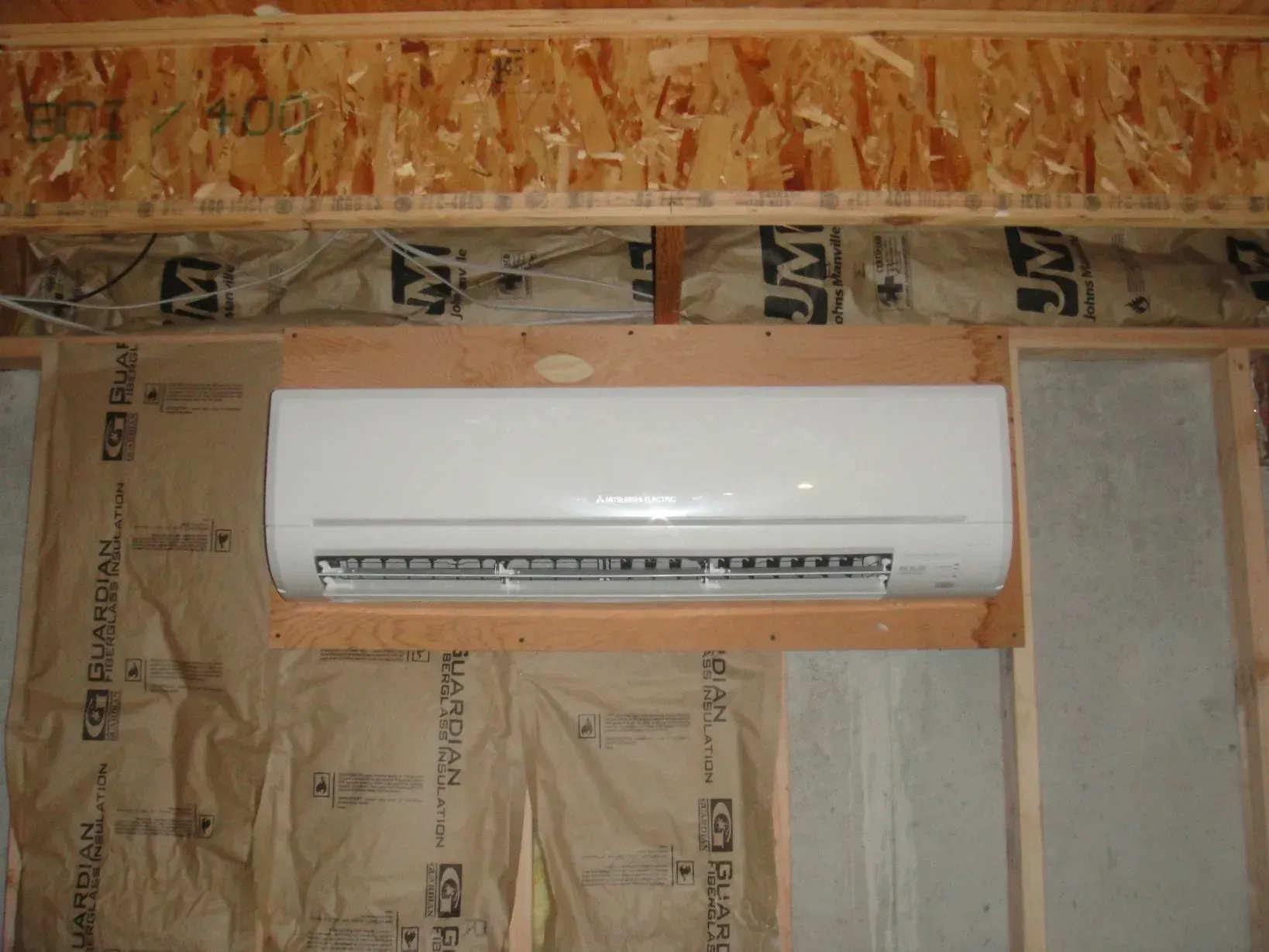 Wall-mounted white air conditioner in an unfinished room with exposed insulation and wood framing