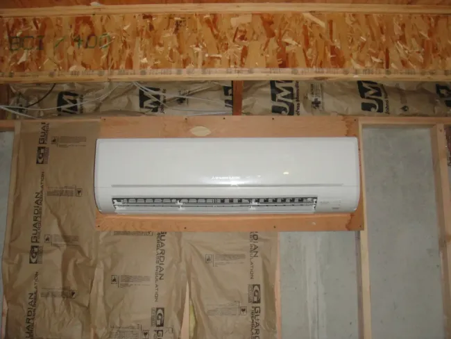 Wall-mounted white air conditioner in an unfinished room with exposed insulation and wood framing