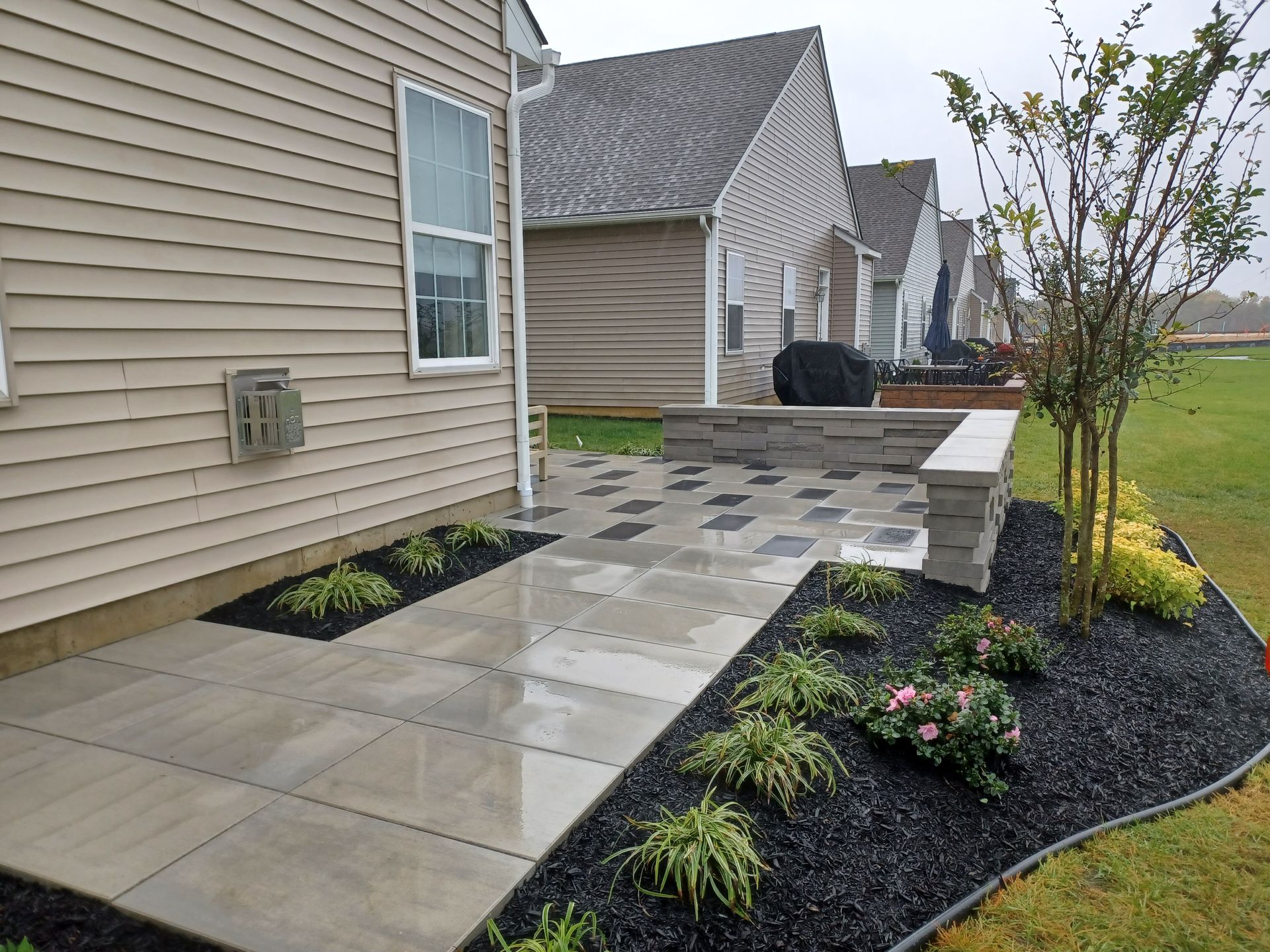 Patio with pavers and landscaping next to a house with beige siding and neighboring houses in the background.
