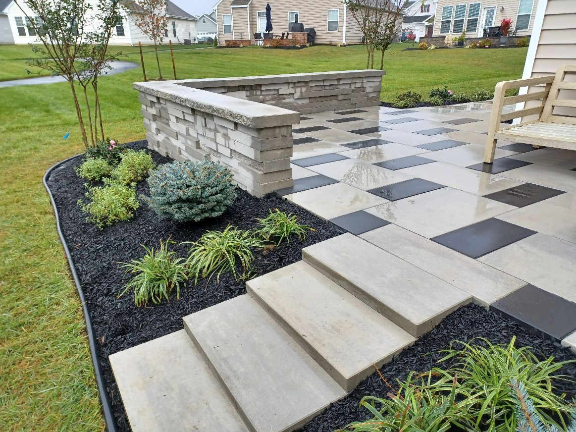 Stone patio with steps and retaining wall, edged with black mulch and greenery, adjacent to grass lawn.