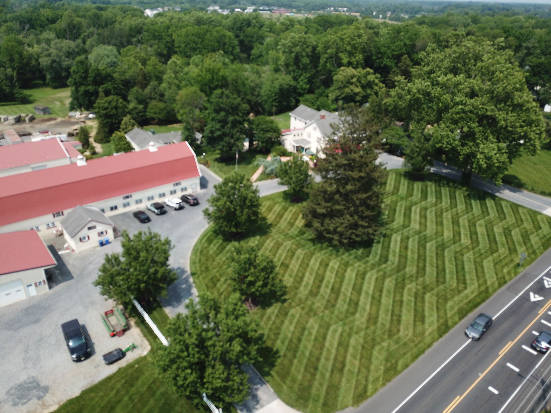 Aerial view: Red-roofed buildings, patterned lawn, trees, road with cars, and a white house on a green landscape.