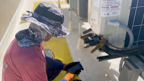 Person cleaning or inspecting a wall-mounted unit in a hallway, wearing a wide-brim hat and gloves.