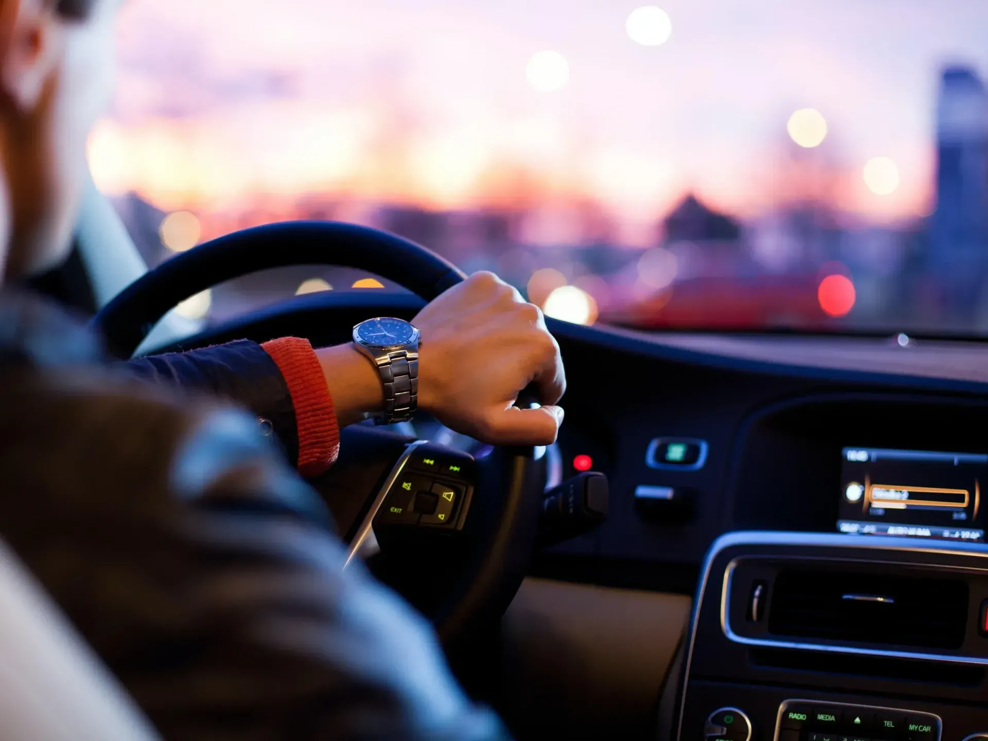 Person driving a car at dusk, hand on the steering wheel, with city lights in the background.