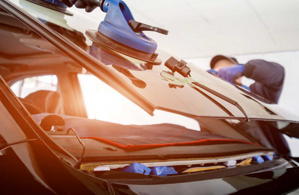 A worker installing a car windshield using suction cups and tools in a shop.