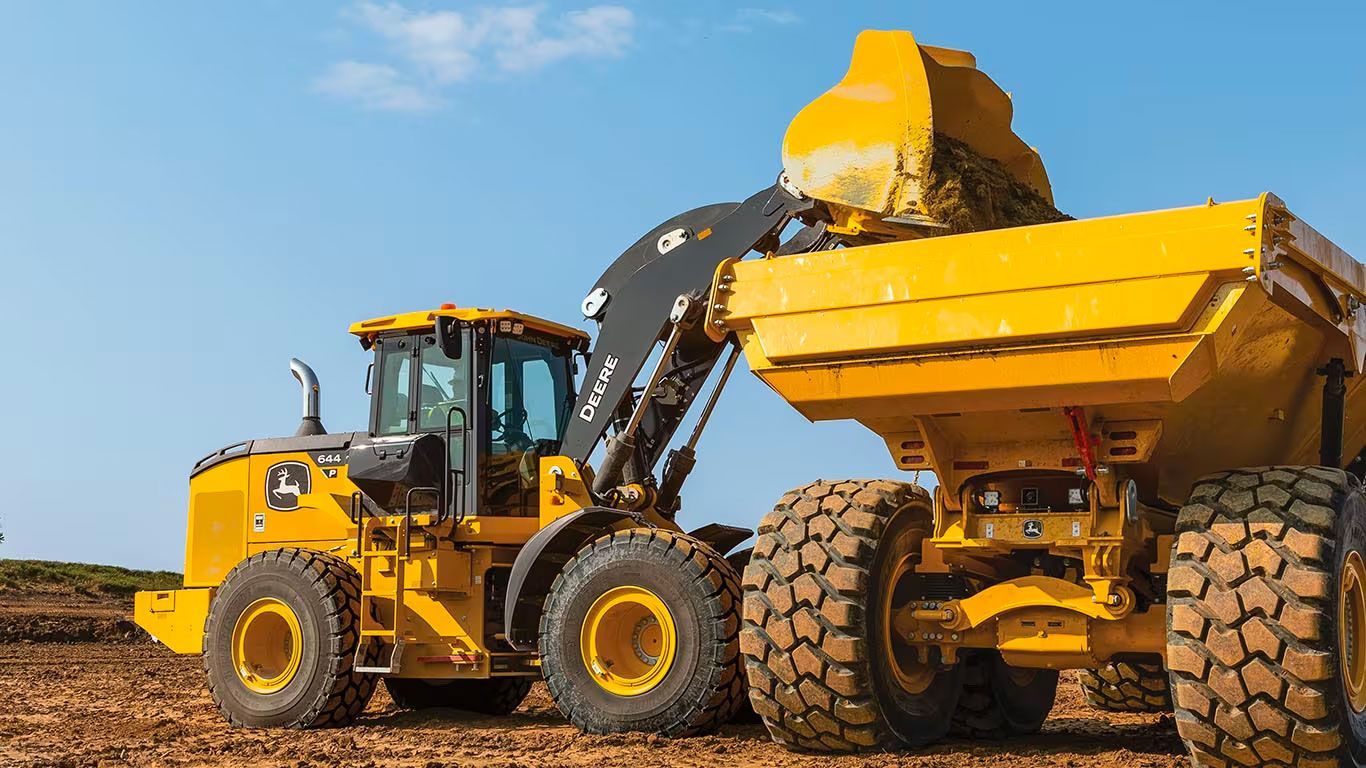 Yellow Deere wheel loader dumping earth into a yellow dump truck on a construction site under a blue sky.