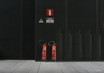 Two red fire extinguishers stand before a black metal panel with safety signs.