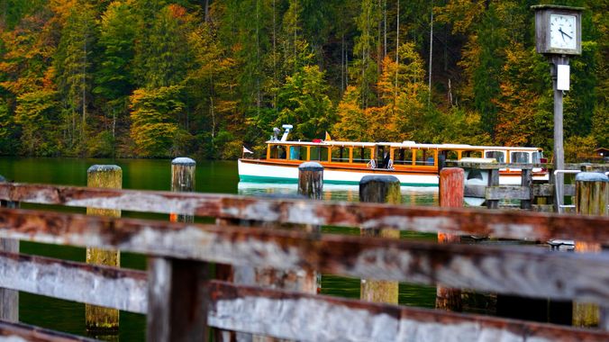 Rainy riverside dock with a moored tour boat, metal railing in foreground, and forested hillside in the background