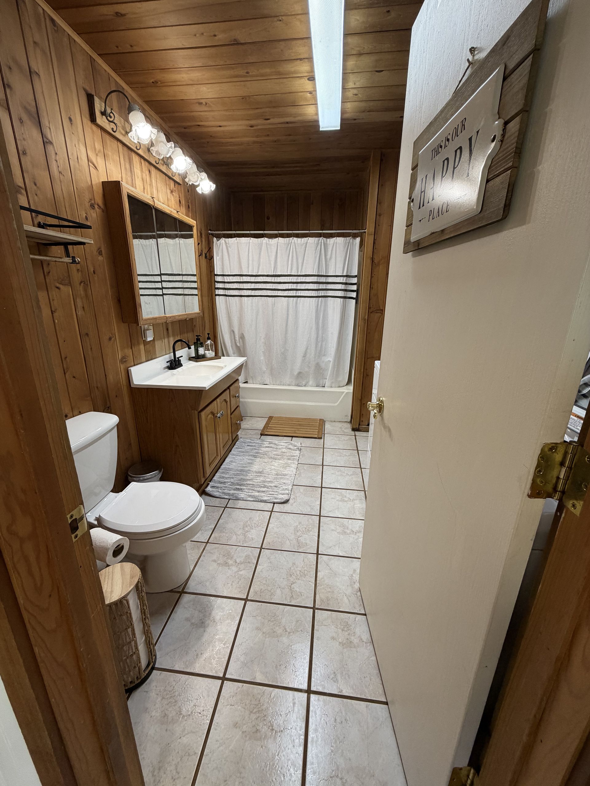 Narrow rustic bathroom with toilet, sink, shower curtain, tiled floor, and wood-paneled walls and ceiling