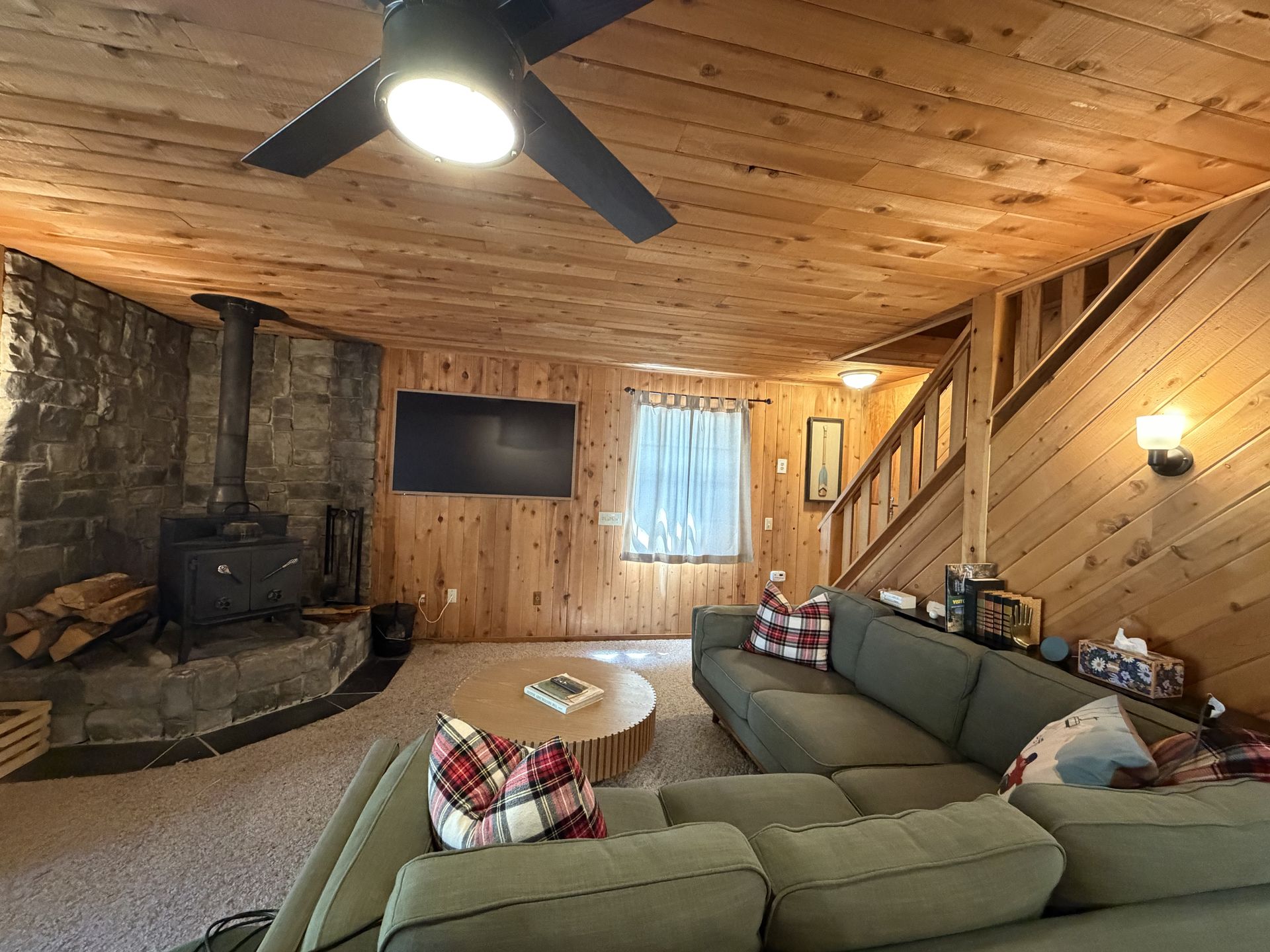 Cozy cabin living room with wood-paneled walls, sectional sofa, stone fireplace, and ceiling fan.