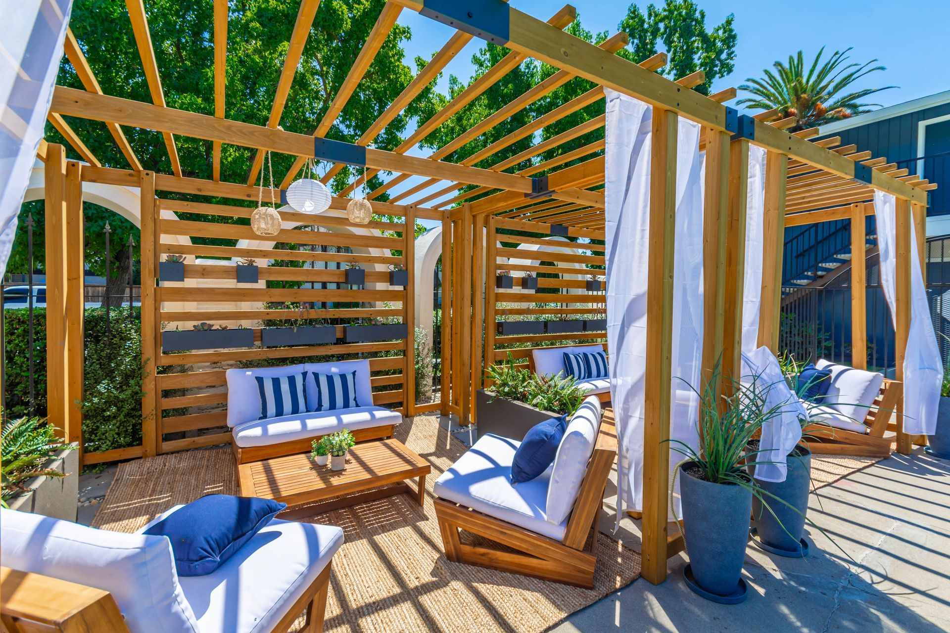 Sunlit cabana lounge with white-and-blue cushioned chairs, striped curtains, and wooden pergola by the pool