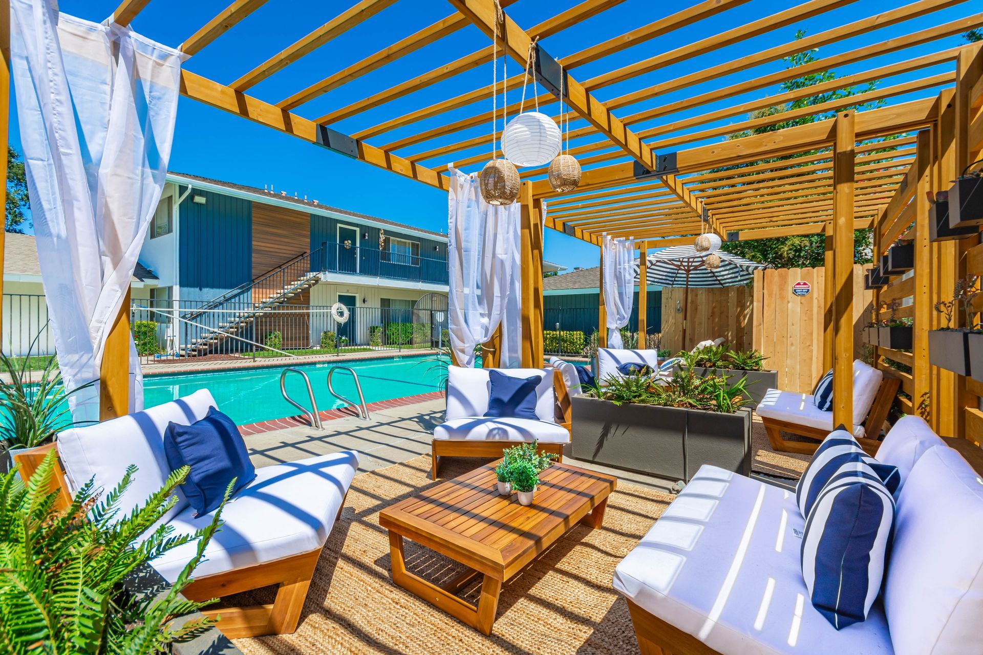 Patio lounge with white-and-blue cushions, a pool, and a wooden pergola in bright sunlight