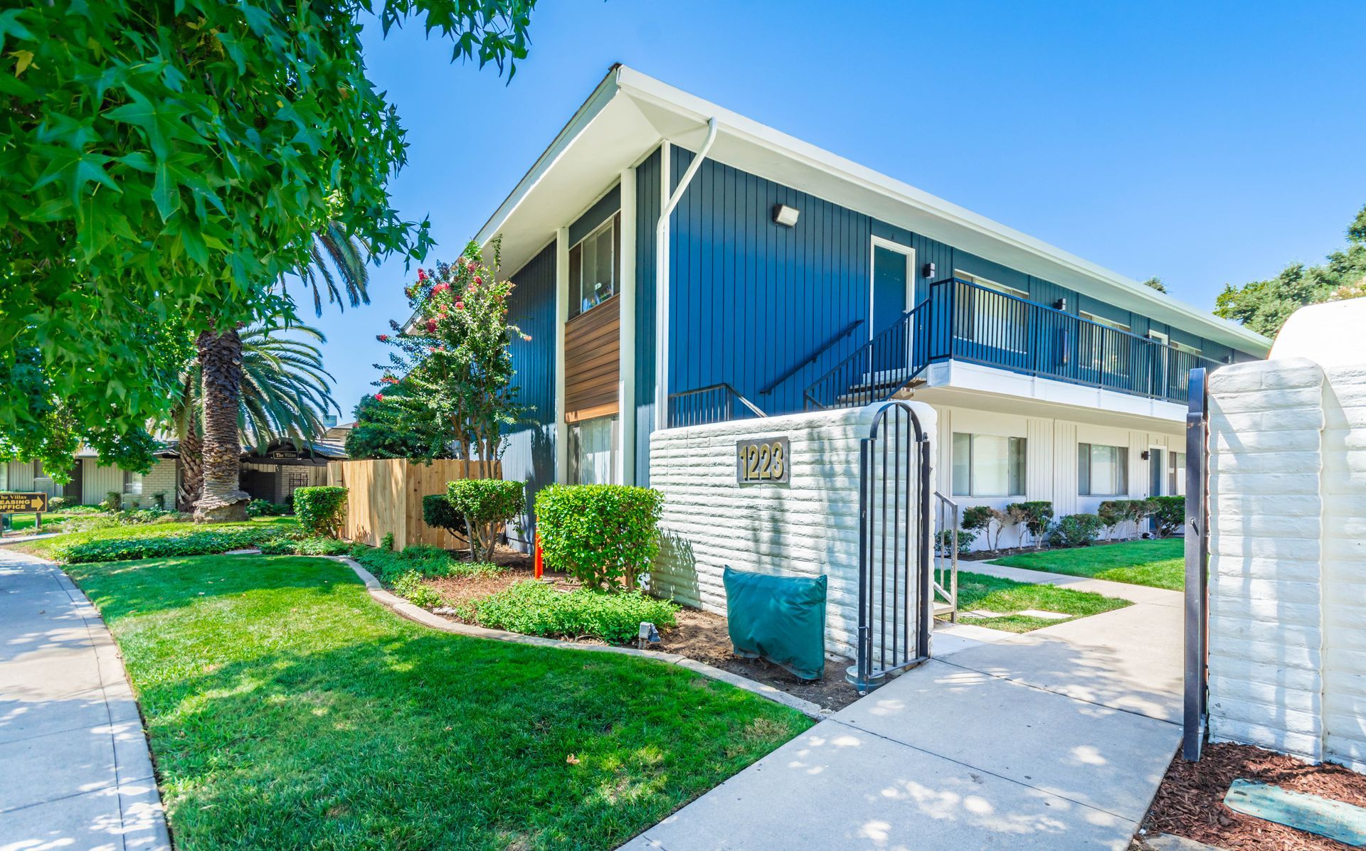 Modern two-story apartment building with blue balcony railings, green lawn, and sidewalk in bright sunlight