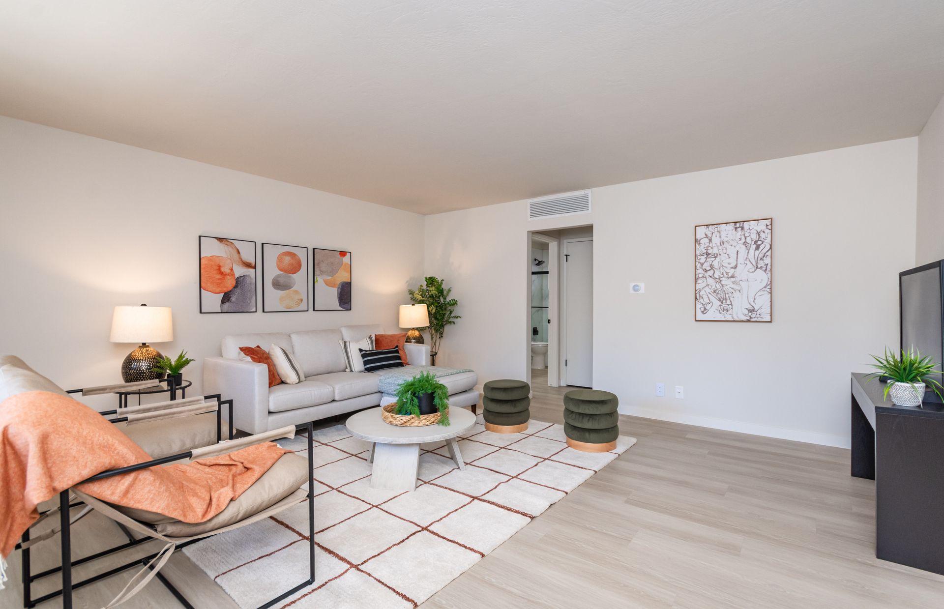 Bright modern living room with white sofa, orange chair, coffee tables, rug, floor lamp, and wall art