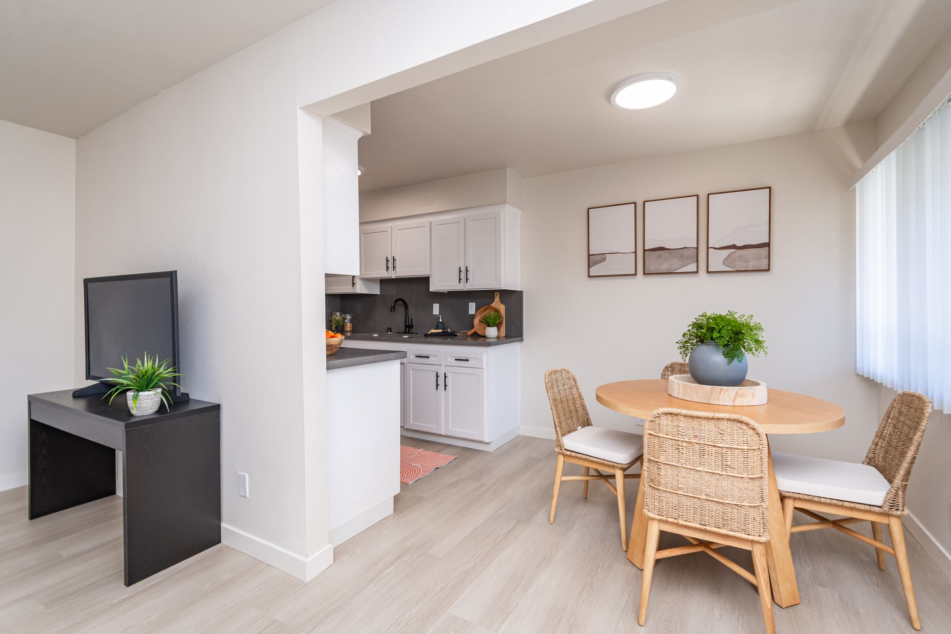 Bright open dining area with wicker chairs, round wooden table, and a small kitchen in the background