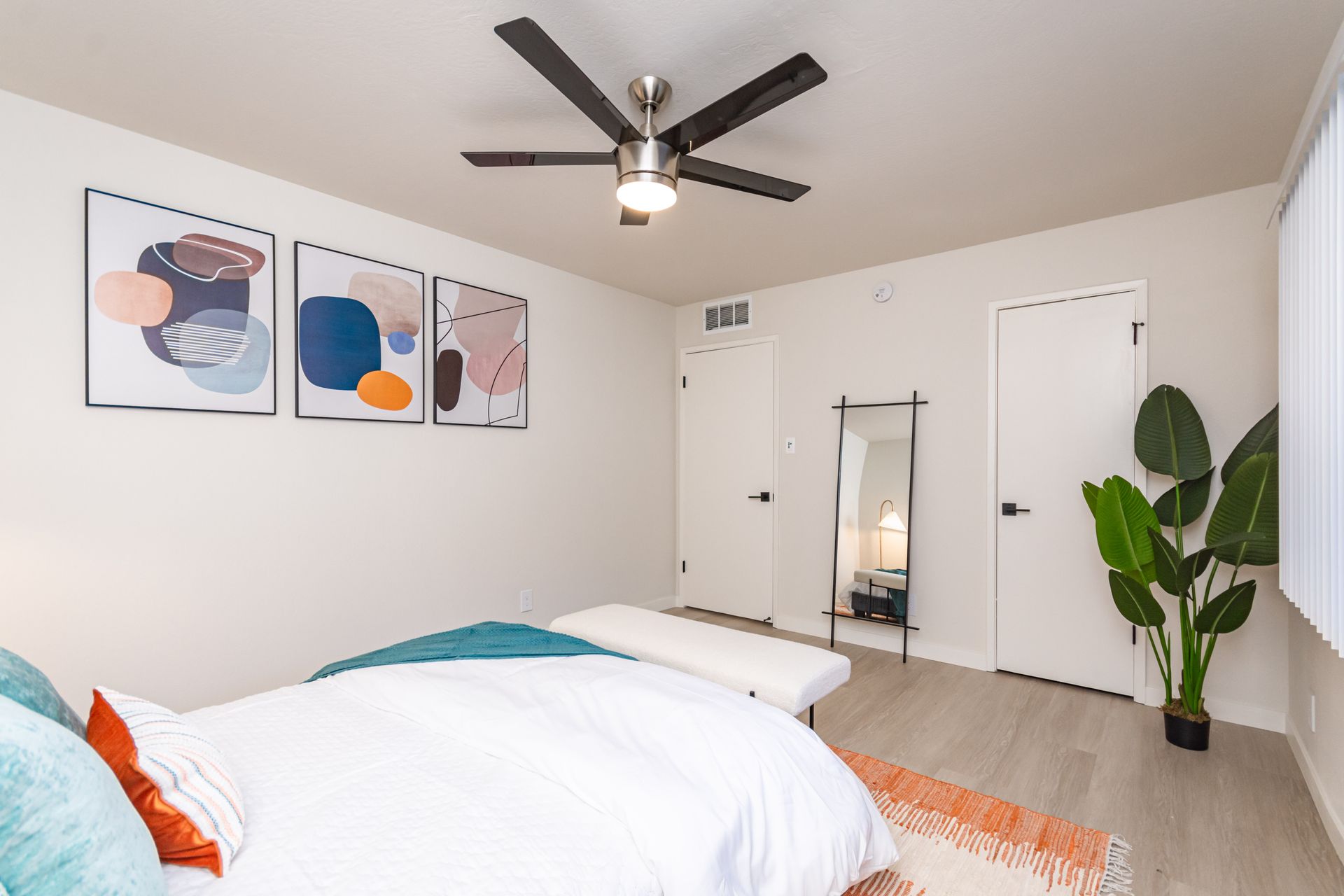 Bright bedroom with white bedding, abstract wall art, ceiling fan, mirror, and a tall green plant