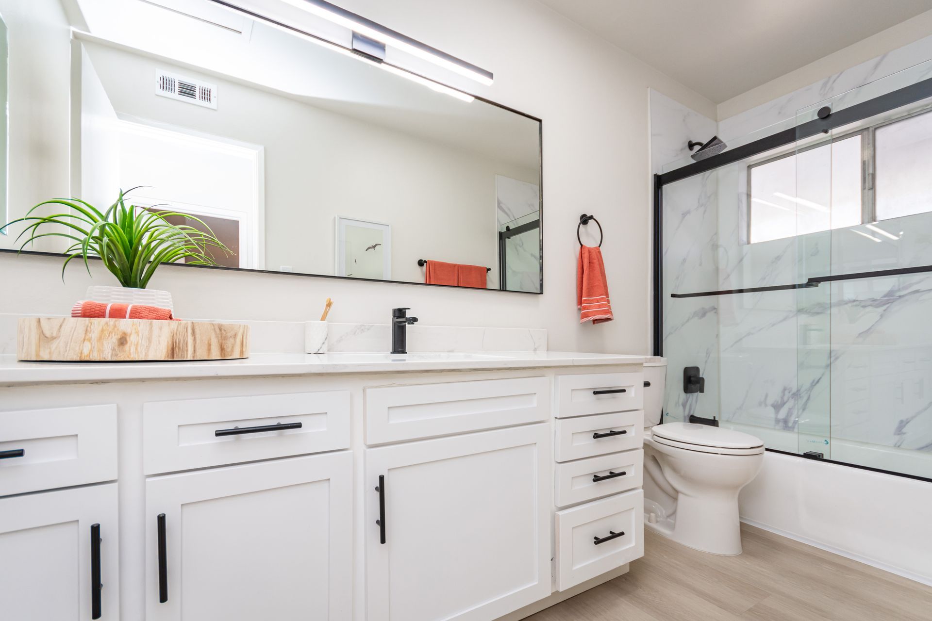 Bright white bathroom with double vanity, large mirror, toilet, shower, and coral towel accent