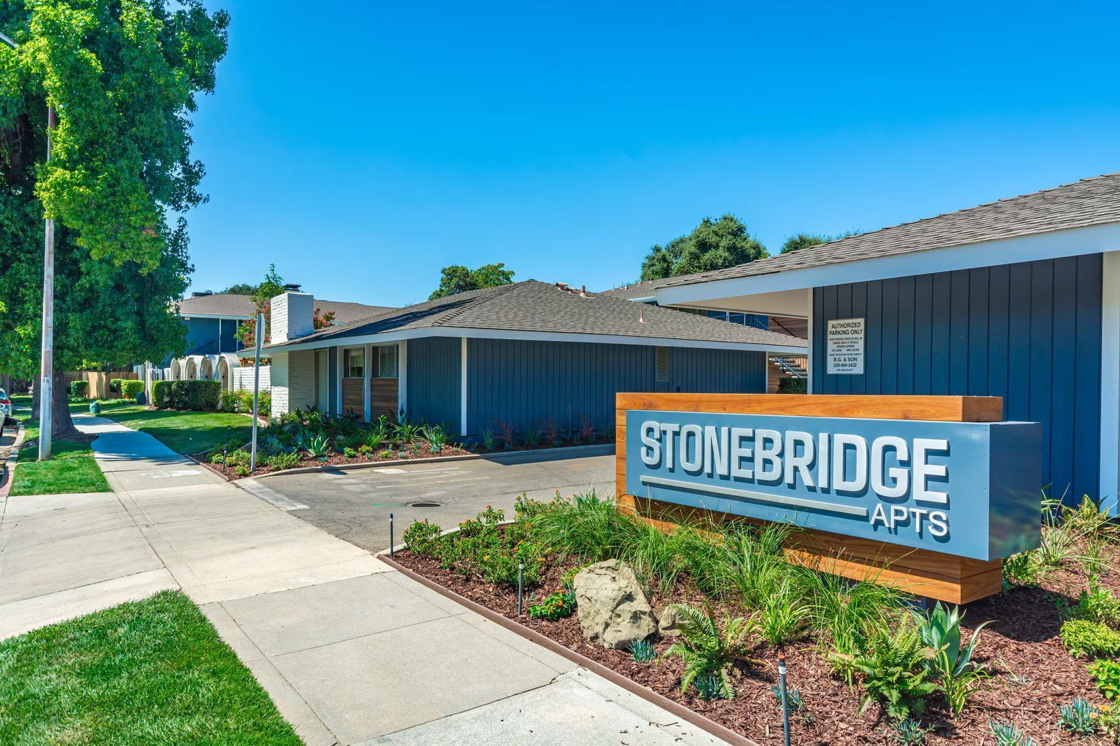 Stonebridge Apartments sign in front of a blue building on a sunny sidewalk, with trees and landscaping.