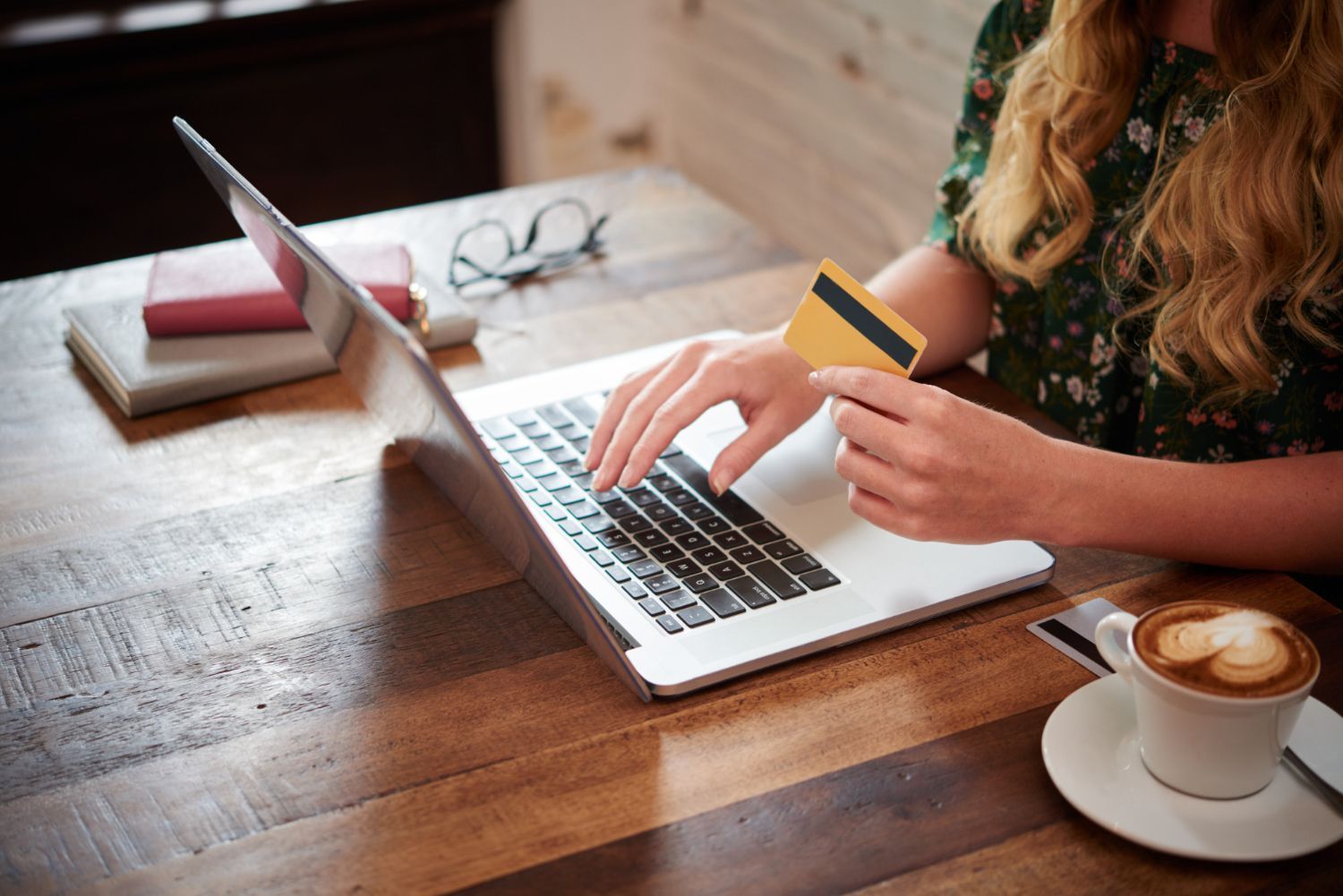 Person typing on a laptop at a wooden desk, holding a credit card beside a cup of coffee.