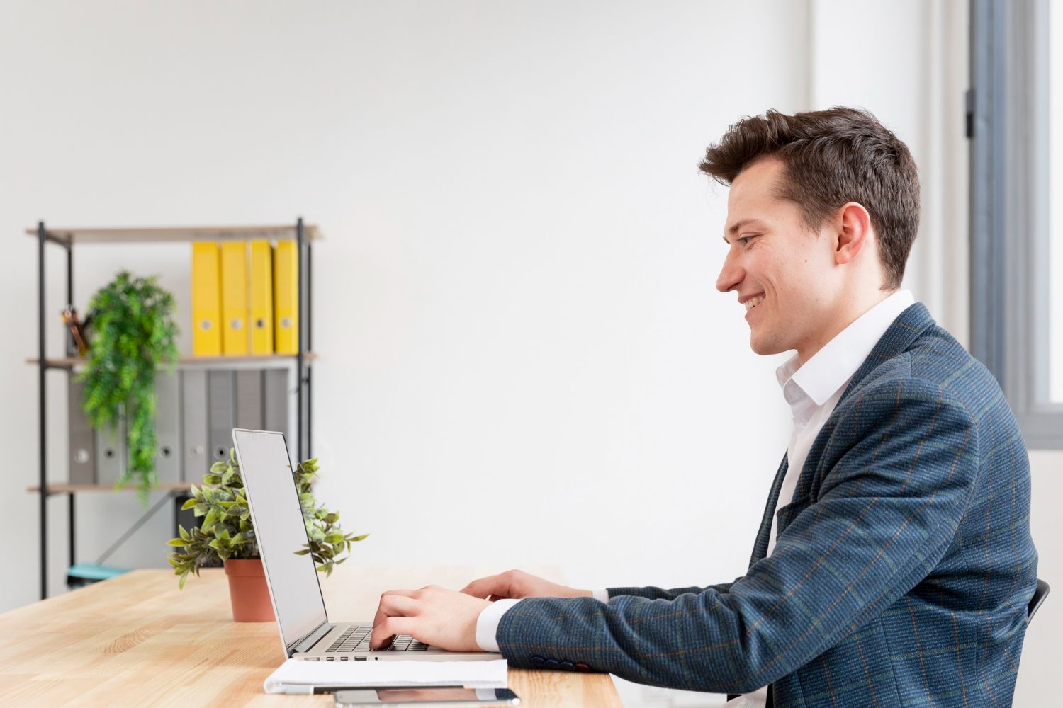 Man using a laptop at a desk in a bright office, smiling while working