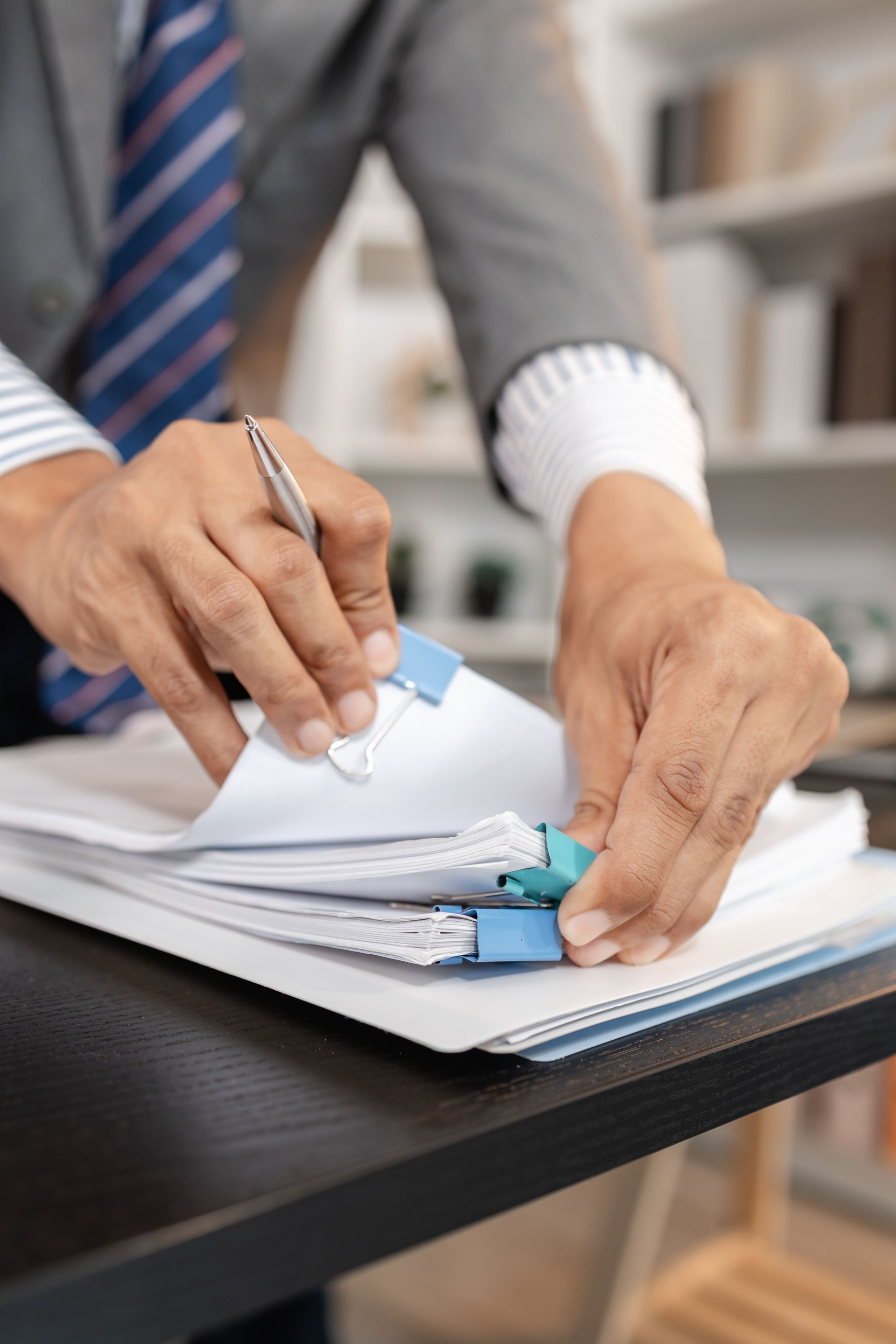 Hands signing documents on a desk with a blue pen and tie visible