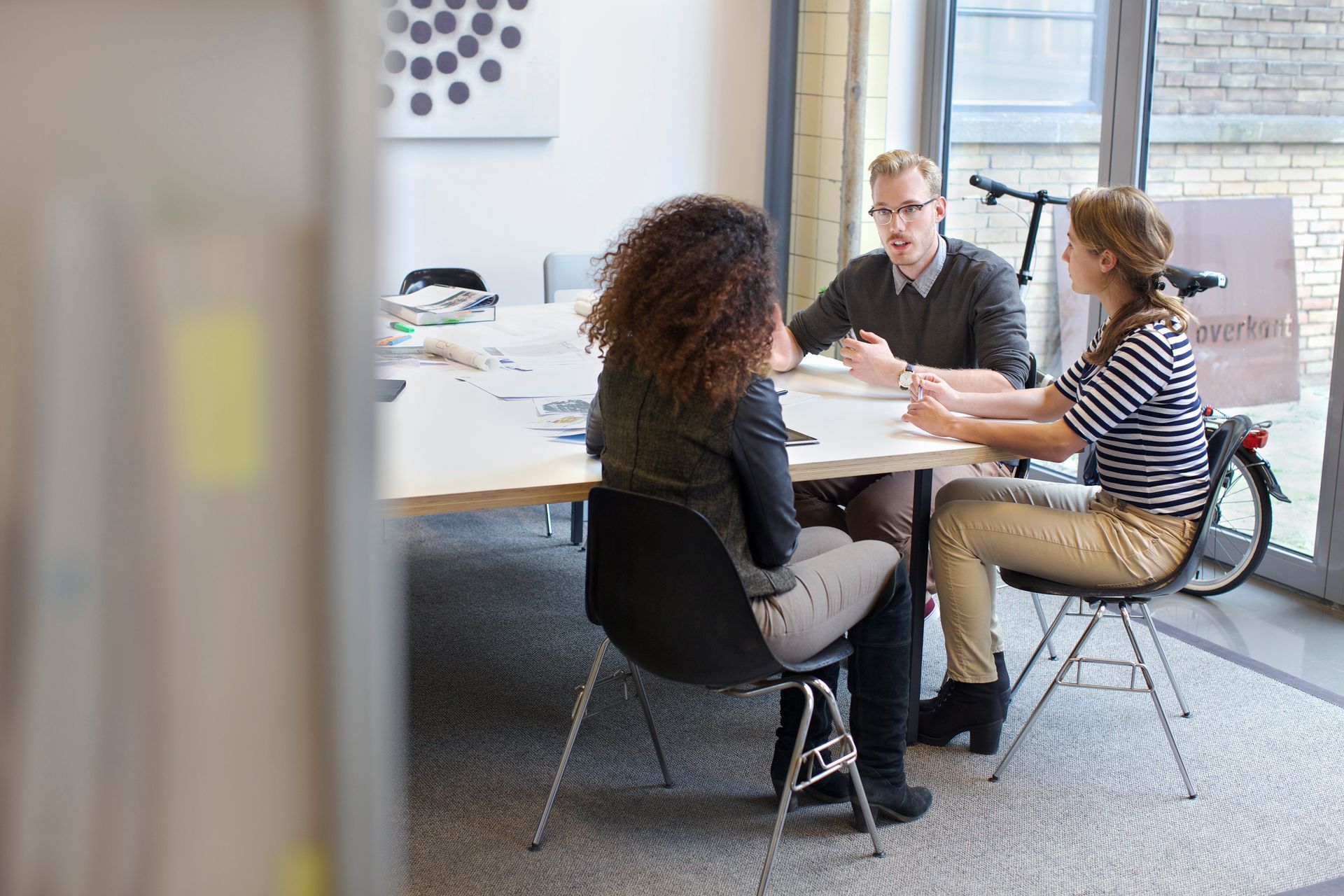 Three people in a bright office meeting around a table, discussing with papers and a laptop.
