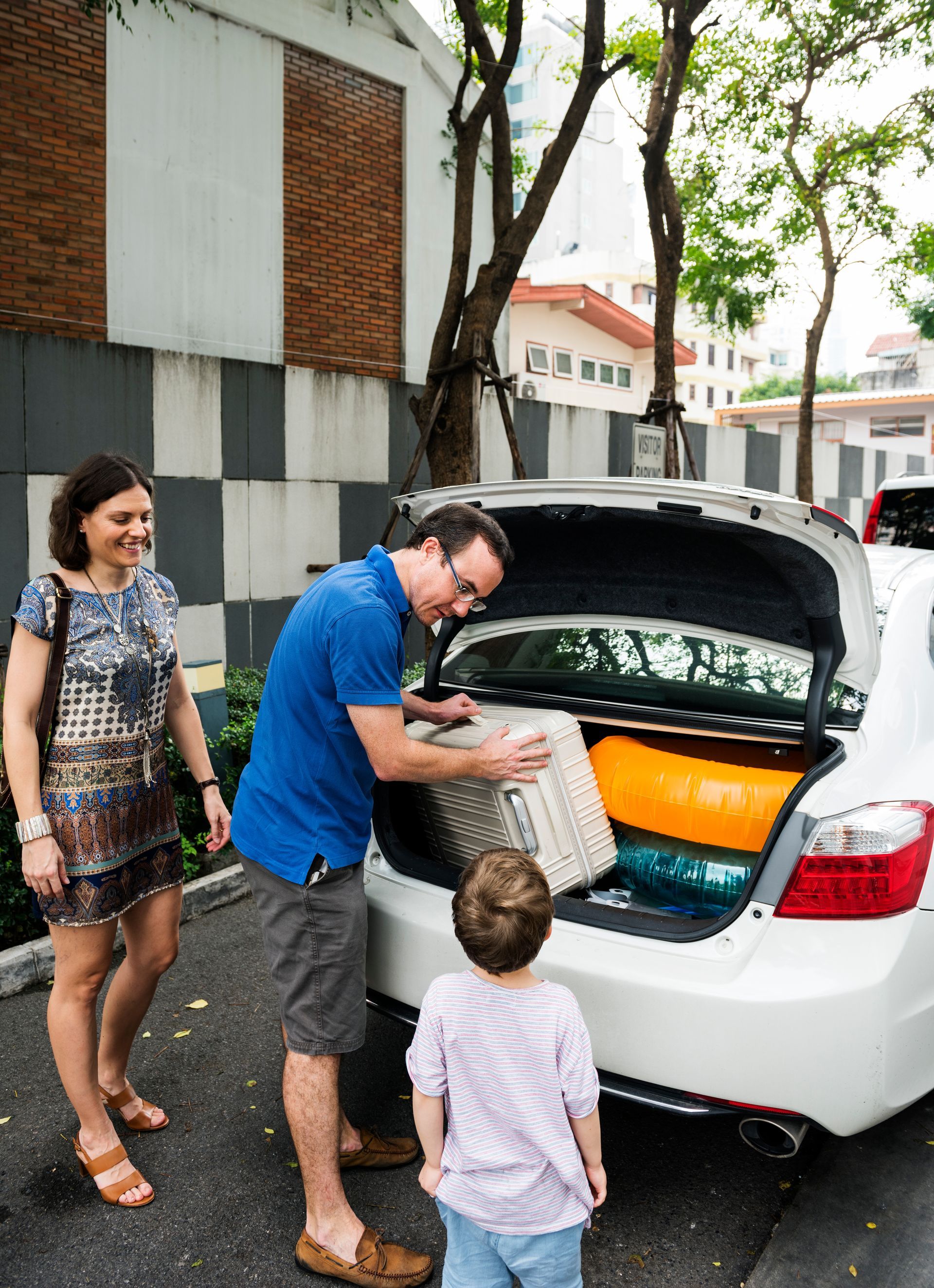 People loading a white car trunk with bags on a tree-lined street