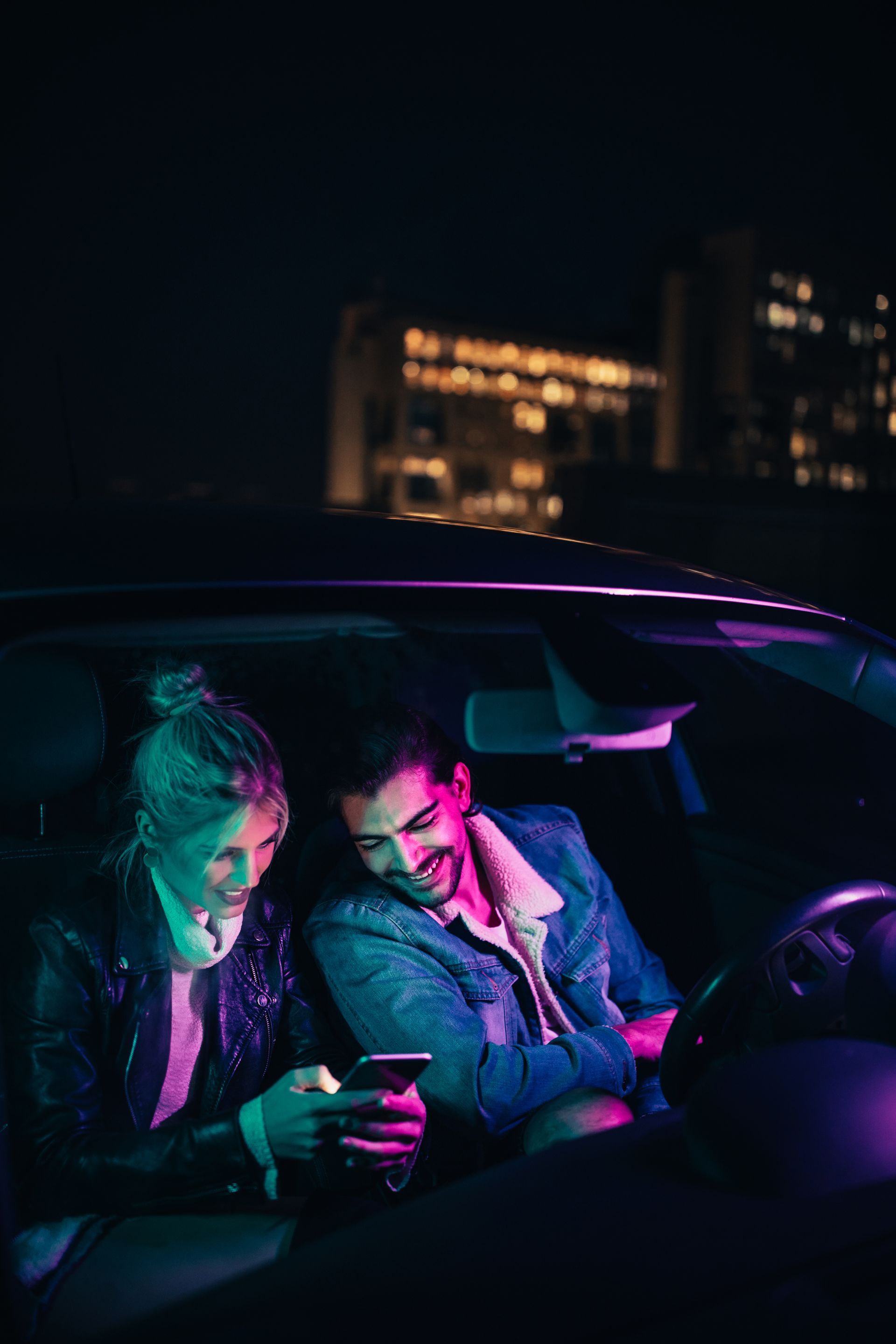 Two people in a car at night, lit by neon pink and teal, looking at a phone.