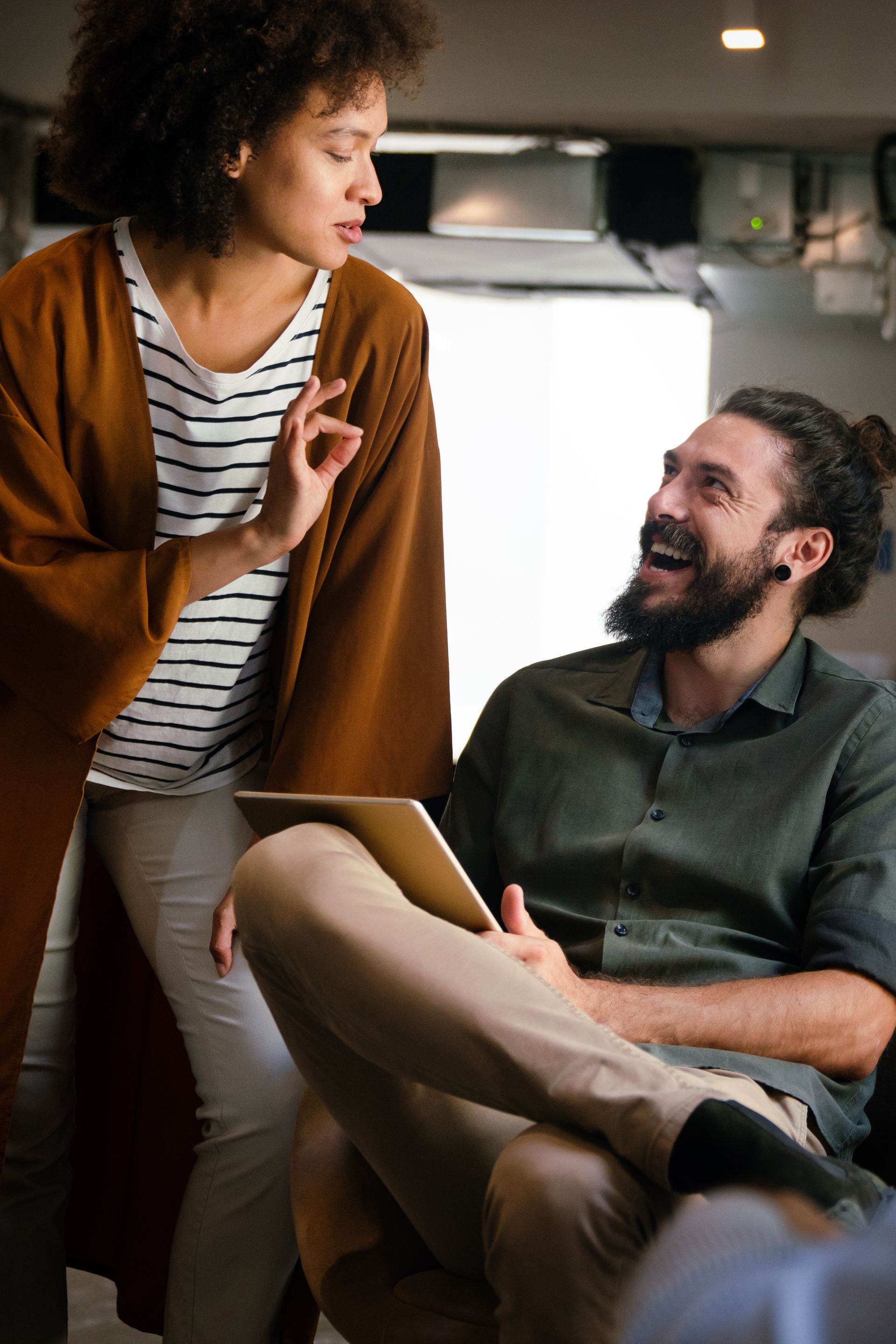 Two coworkers chatting in a kitchen, one seated with a laptop and the other standing and gesturing.
